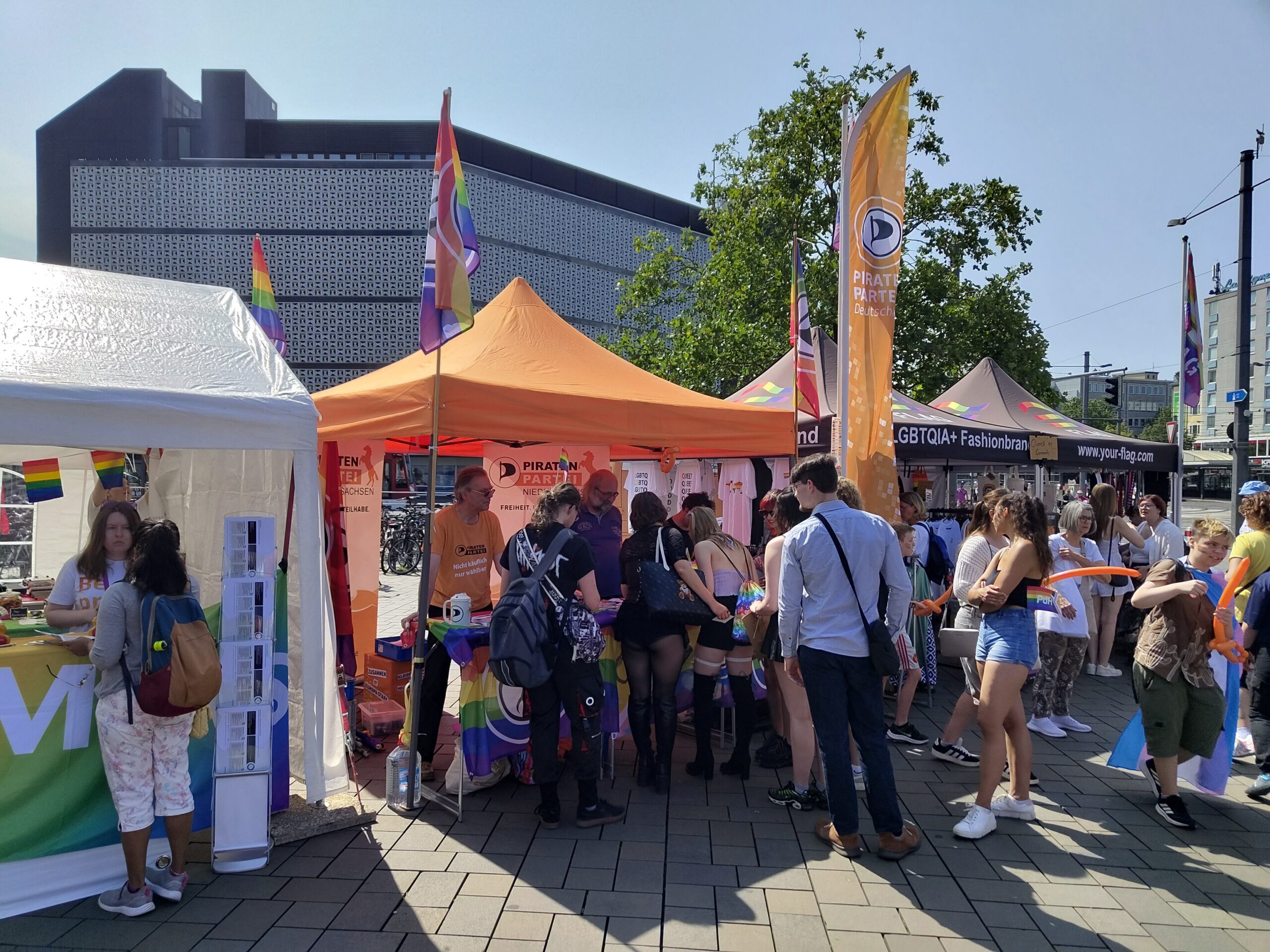 Stand der Piratenpartei mit Baldachin. An den Ecken des Standes unterschiedliche Piratenflaggen. Vor dem Stand, welcher mit weiteren Flaggen geschmückt ist, stehen Personen und betrachten die angebotenen Sticker. Dahinter mehrere Mitglieder der Piratenpartei Niedersachsen. Seitlich einige Personen mit orangen Luftballon-Piratensäbeln. 