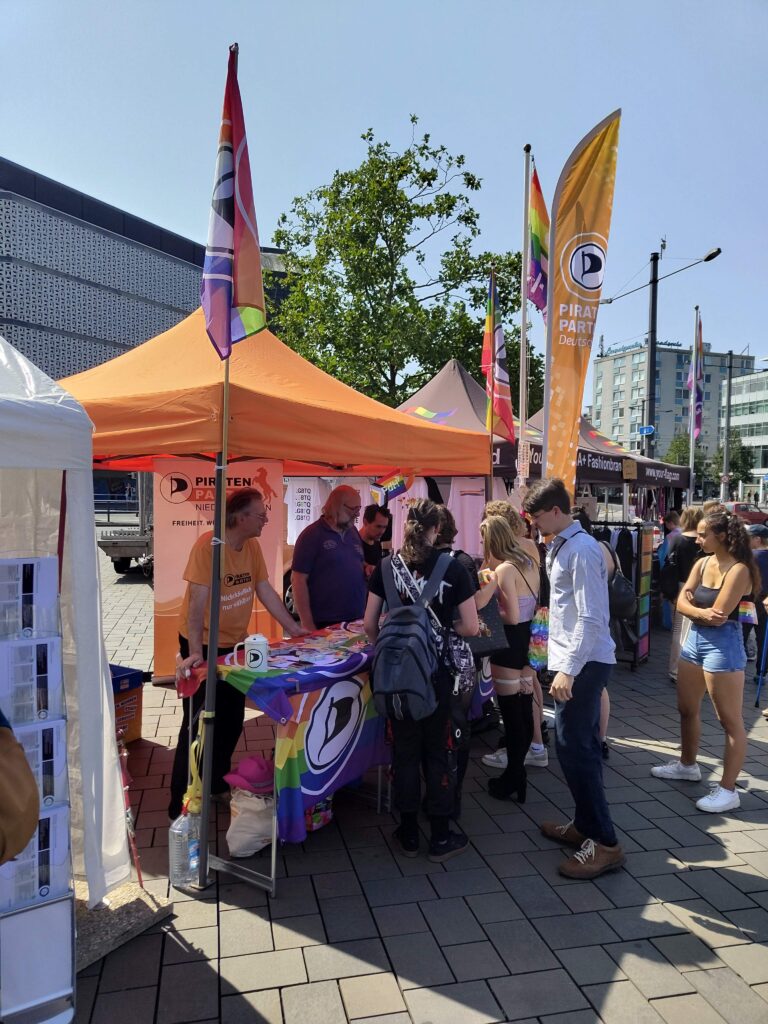 Stand der Piratenpartei mit Baldachin. An den Ecken des Standes unterschiedliche Piratenflaggen. Vor dem Stand, welcher mit weiteren Flaggen geschmückt ist, stehen Personen und betrachten die angebotenen Sticker. Dahinter mehrere Mitglieder der Piratenpartei Niedersachsen.