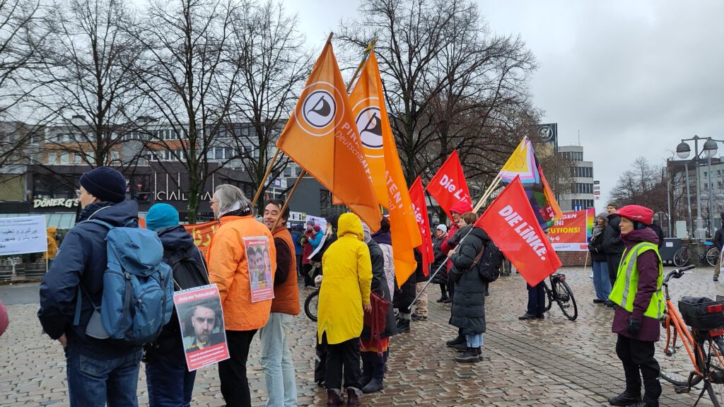 Menschen stehen im Kreis; einige halten Flaggen in ihren Händen: Piraten und Die Linke.