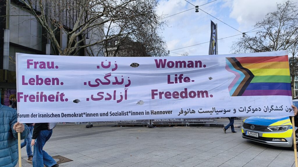 Banner, welches von zwei Personen gehalten wird: Der Slogan 'Frau. Leben. Freiheit' in deutscher, persischer und englischer Sprache. Daneben die Progress-Flagge. Forum der iranischen Demokrat*innen und Sozialst*innen in Hannover.
