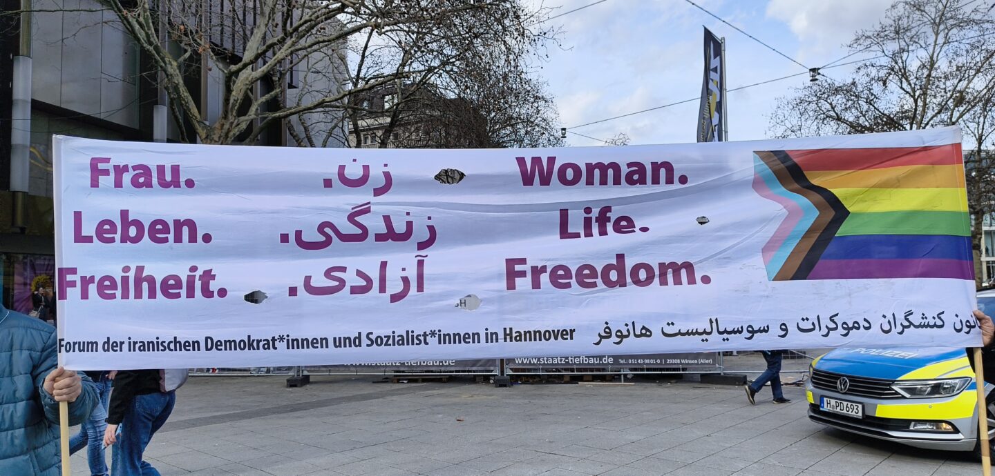 Banner, welches von zwei Personen gehalten wird: Der Slogan 'Frau. Leben. Freiheit' in deutscher, persischer und englischer Sprache. Daneben die Progress-Flagge. Forum der iranischen Demokrat*innen und Sozialst*innen in Hannover.