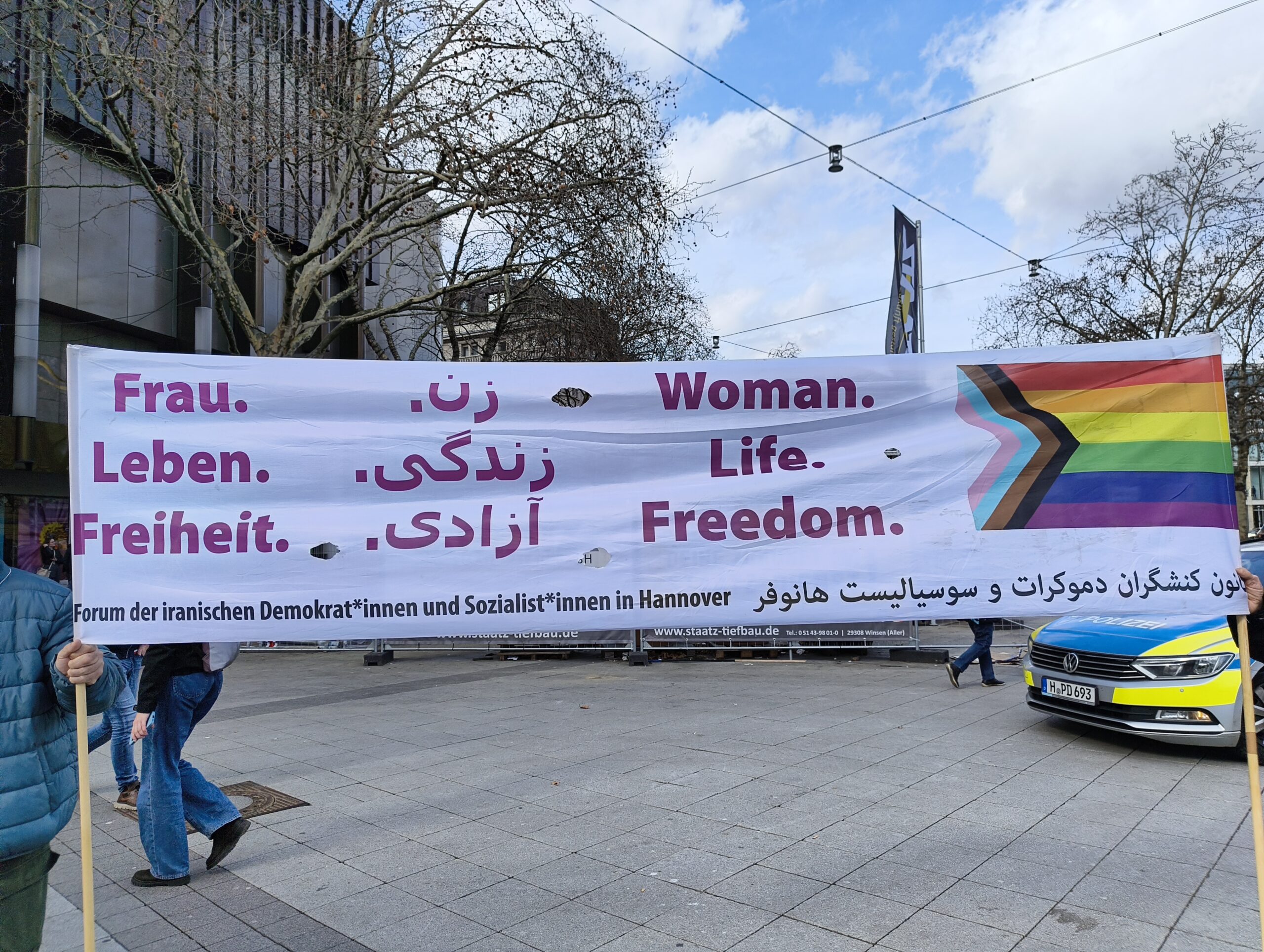 Banner, welches von zwei Personen gehalten wird: Der Slogan 'Frau. Leben. Freiheit' in deutscher, persischer und englischer Sprache. Daneben die Progress-Flagge. Forum der iranischen Demokrat*innen und Sozialst*innen in Hannover.