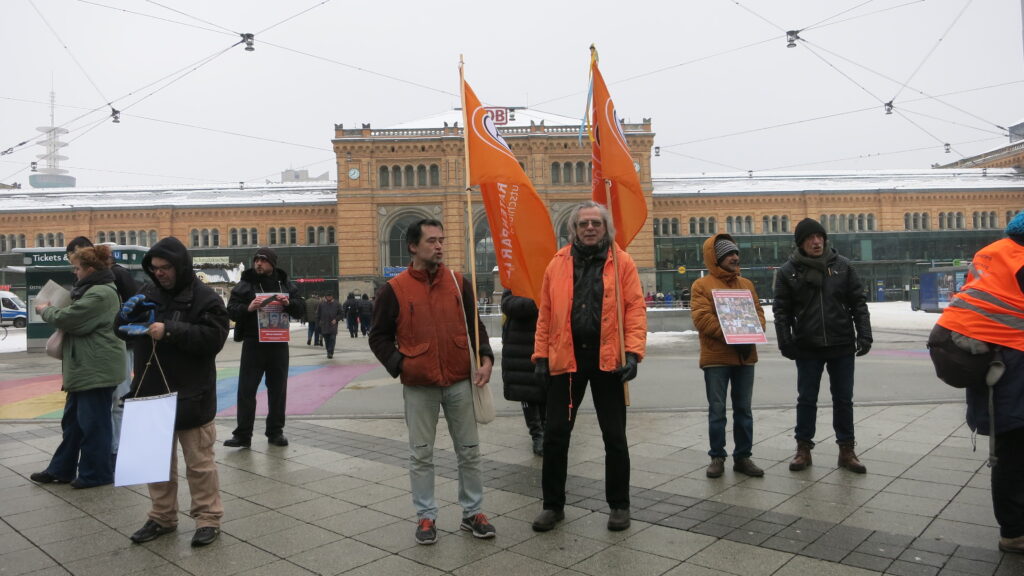 Zwei Personen mit Fahnen der Piratenpartei Deutschlands stehen mit weiteren Personen vor dem Hauptbahnhof Hannover.
