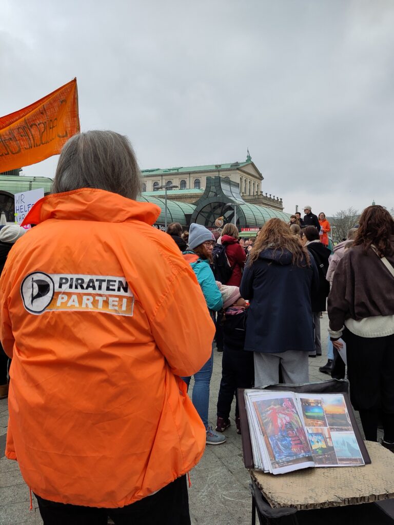 Blick in die Menschenmenge; die Menschen drehen dem Betrachtenden den Rücken zu. Eine der Personen im Vordergrund trägt eine orange Jacke mit dem Logo der Piratenpartei.