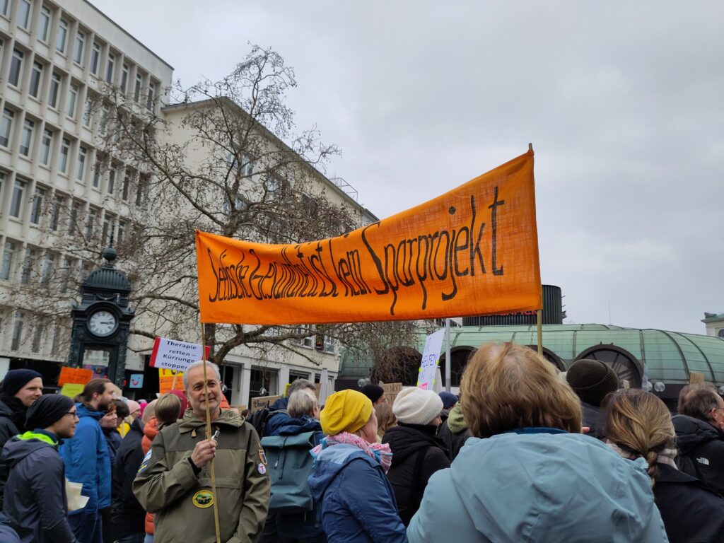 Ein Banner wird von zwei Personen an Stangen gehalten. Auf dem Banner der Text „Seelische Gesundheit ist kein Sparprojekt“.