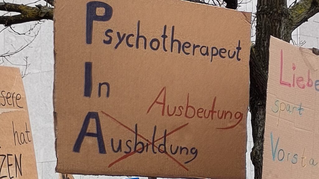 Handschriftliches Plakat mit dem Text „Psychotherapeut In Ausbildung (PIA)“ – das Wort Ausbildung ist ausgekreuzt und durch das Wort „Ausbeutung“ ersetzt.