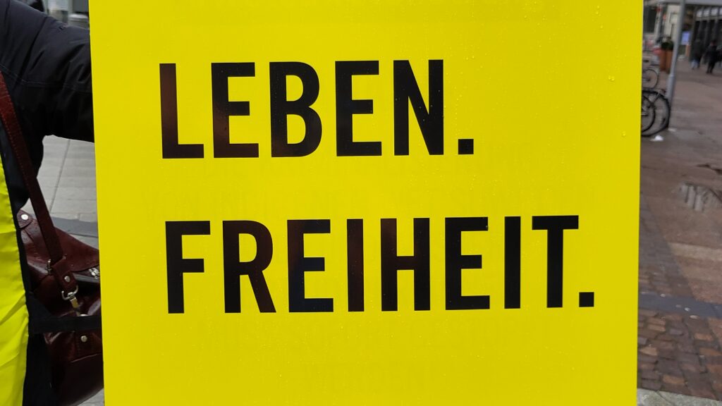 Schild mit dem Text „Frau. Leben. Freiheit“ mit schwarzem Text auf gelbem Hintergrund. Darunter das Logo von Amnesty International. Eine Hand hält das Schild am oberen Ende fest.