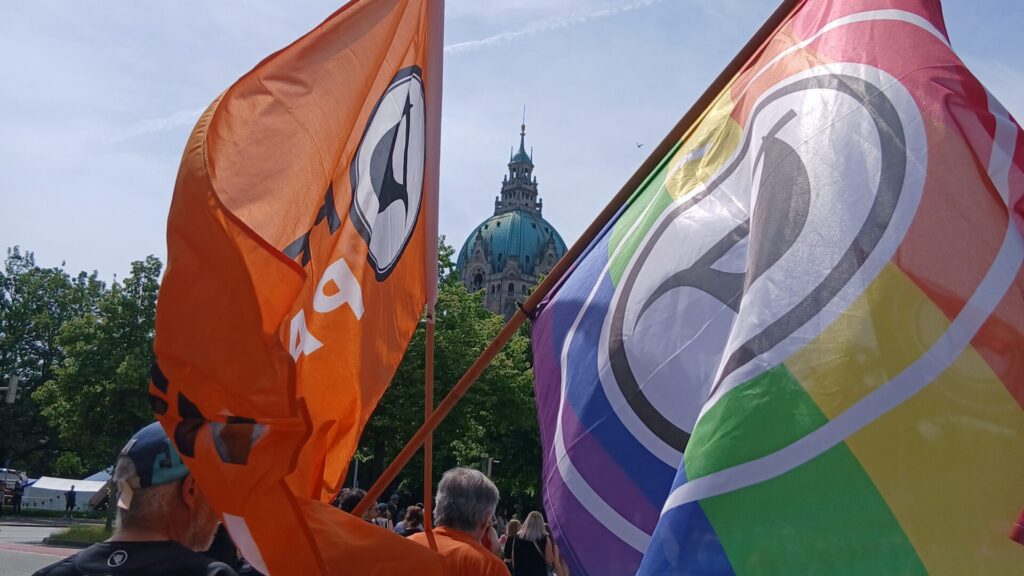 Personen auf einer Demo. Im Mittelpunkt zwei Flaggen der Piratenpartei, eine davon mit Regenbogenstreifen. In der Mitte, zwischen den Flaggen, die Kuppel des Neuen Rathauses.
