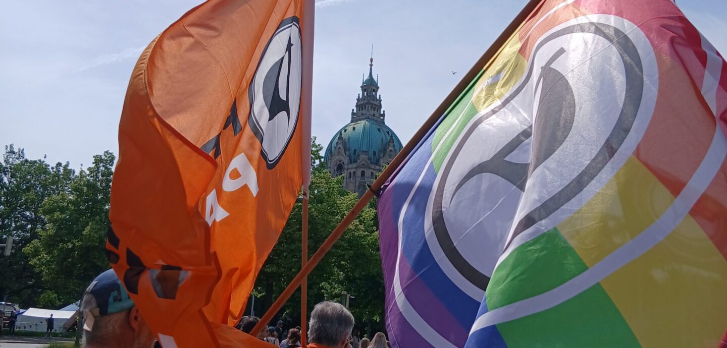 Personen auf einer Demo. Im Mittelpunkt zwei Flaggen der Piratenpartei, eine davon mit Regenbogenstreifen. In der Mitte, zwischen den Flaggen, die Kuppel des Neuen Rathauses.