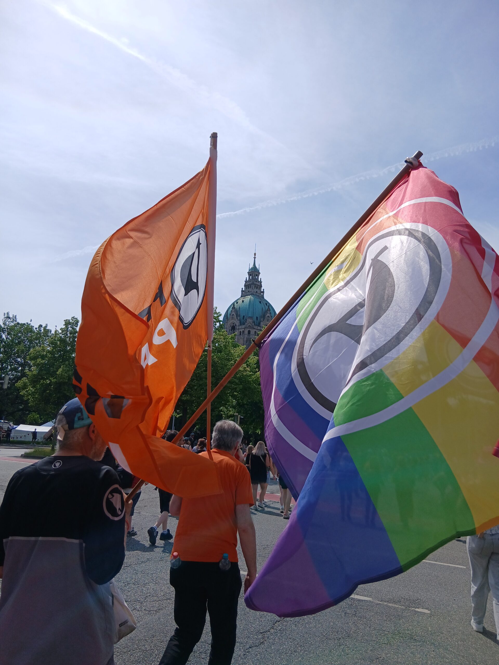 Personen auf einer Demo. Im Mittelpunkt zwei Flaggen der Piratenpartei, eine davon mit Regenbogenstreifen. In der Mitte, zwischen den Flaggen, die Kuppel des Neuen Rathauses.