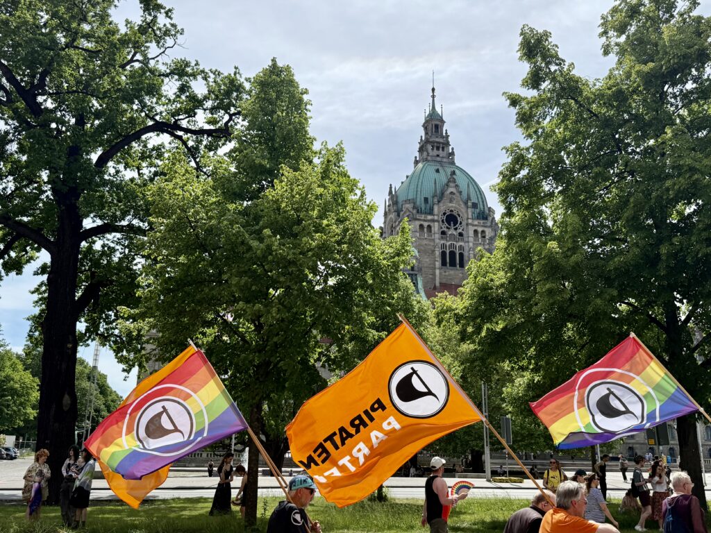 Demoumzug mit verschiedenen Piratenflaggen am unteren Rand des Foto; zwei der Flaggen haben den Regenbogen als Grundmuster. Der Demoumzug geht entlang einer Wiese an grünen Böumen vorbei. Im Hntergrund freie Sicht auf den Turm des Neues Rathauses.