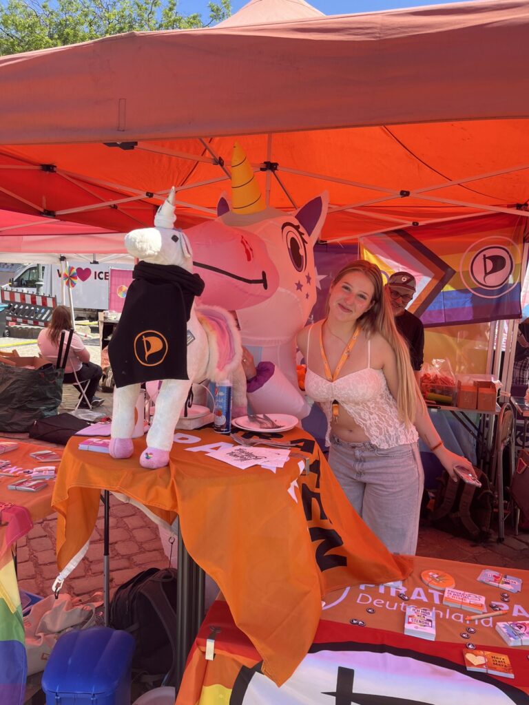 Kayra steht am Stand der Piratenpartei unter einem orangen Baldachin. Sie steht neben einer Person in einem aufblasbaren Einhorn-Kostüm sowie hinter Tischen mit Piratenflaggen, auf denen diverse Sticker liegen. Auf einem runden Stehtisch ein weiteres Einhorn mit einem schwarzen Schal, auf dem das Logo der Piratenpartei deutlich zu erkennen ist.