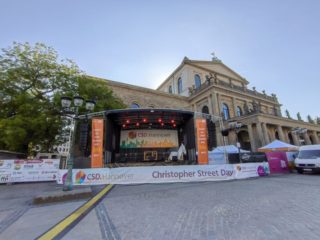 Blick auf die Bühne des CSD.Hannover, die noch unbenutzt ist. Rechts und links an der Bühne Banner mit dem Text »Damit du sein kannst wie du bist.« Im Hintergrund das imponierende Gebäude der Oper, auf der die Regenbogenflagge gehisst ist.