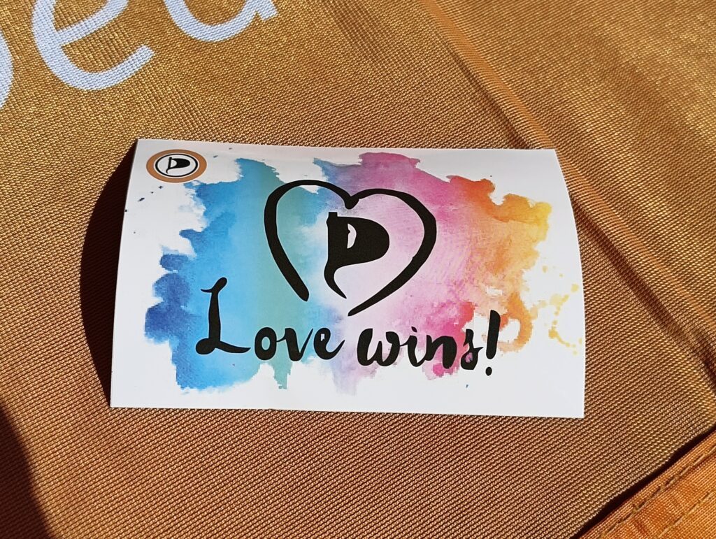 Sticker auf einem orangem Hintergrund: Farbklekse, die ineinander gehen. Darum das Signet der Piratenpartei in einem Herzen. Darunter der Text »Love wins!«