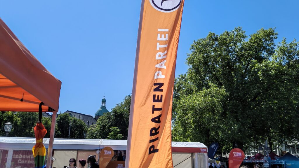 Piratenlogo und Piratenpartei-Schriftzug auf einer Beach-Flagge, in der Nähe eines Standes mit orangem Baldachin steht. Am strahlend blaumen Himmel ist ein Teil der Sonne zu entdecken.
