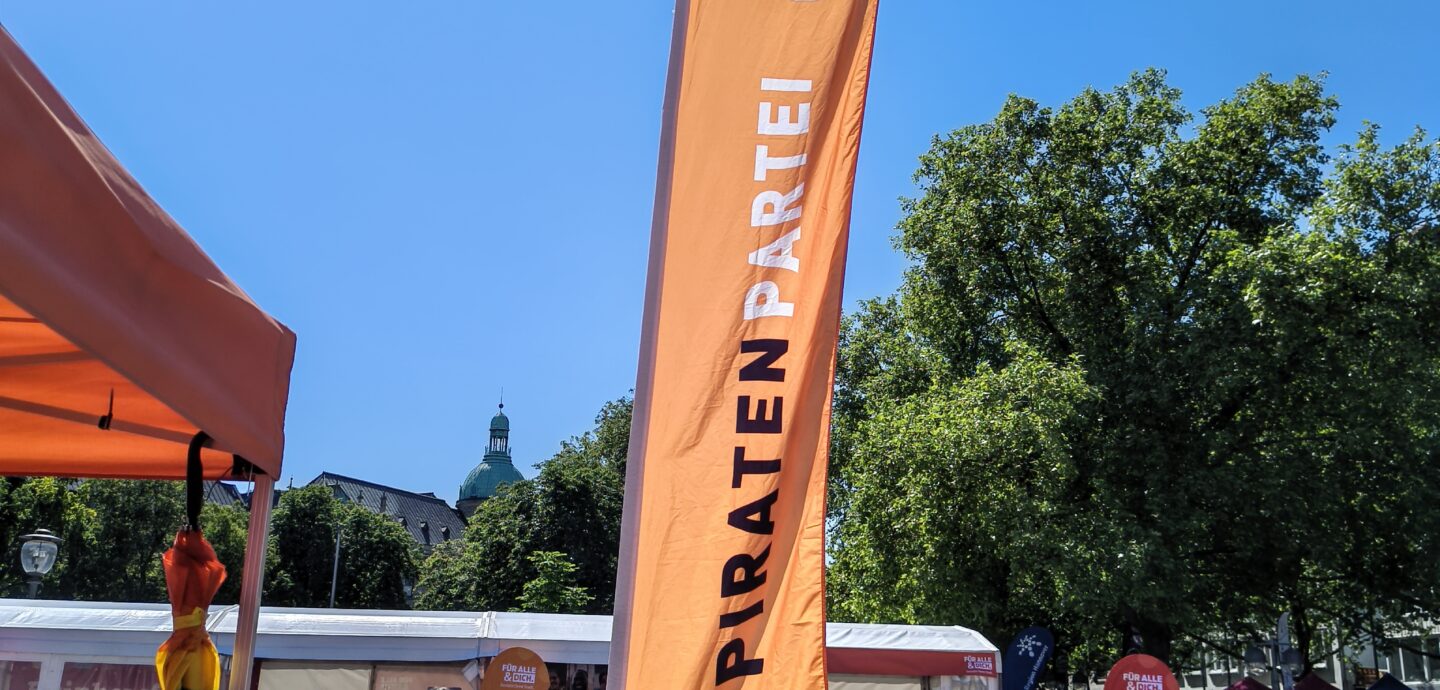 Piratenlogo und Piratenpartei-Schriftzug auf einer Beach-Flagge, in der Nähe eines Standes mit orangem Baldachin steht. Am strahlend blaumen Himmel ist ein Teil der Sonne zu entdecken.