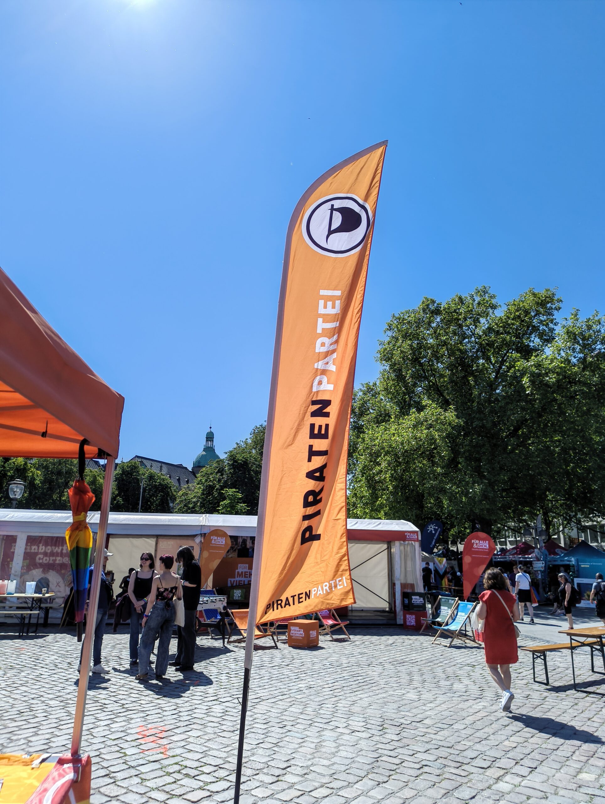 Piratenlogo und Piratenpartei-Schriftzug auf einer Beach-Flagge, in der Nähe eines Standes mit orangem Baldachin steht. Am strahlend blaumen Himmel ist ein Teil der Sonne zu entdecken.