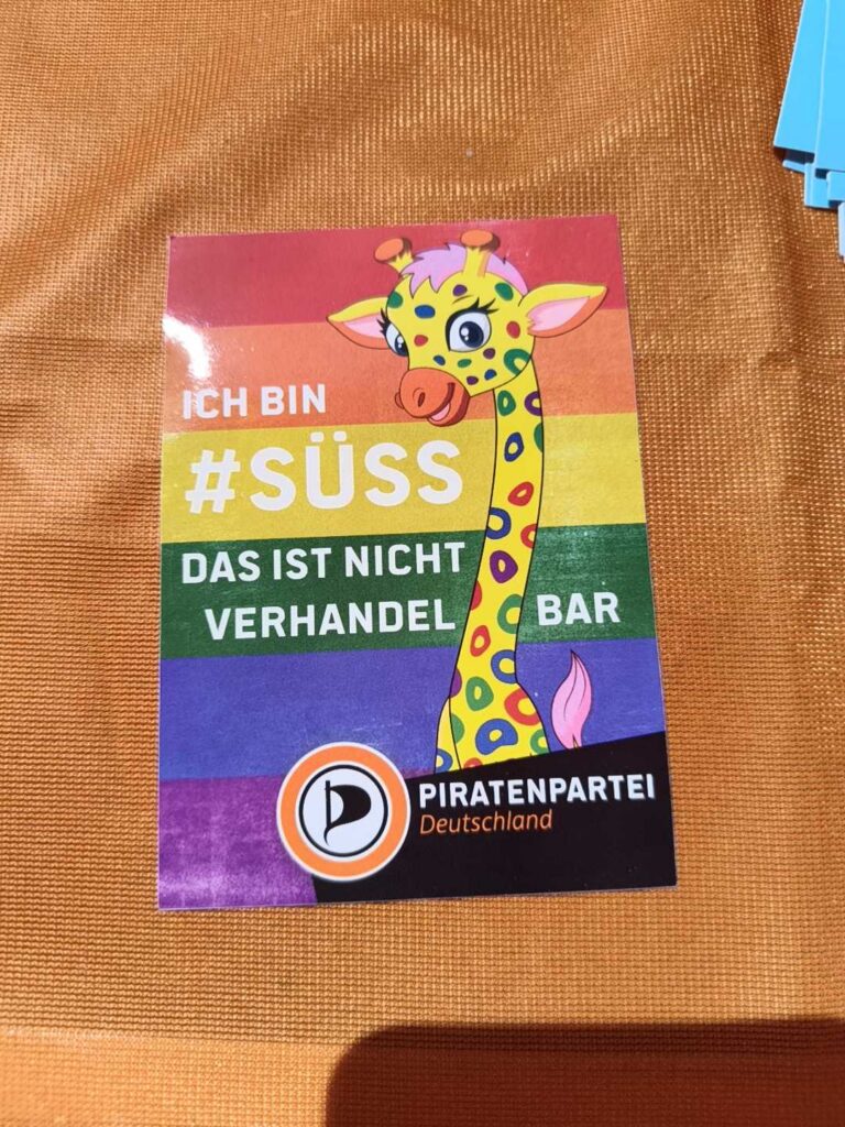 Aufkleber auf einem orangem Hintergrund: Eine Giraffe mit bunten Flecken im Mittelpunkt. Dazu der Text »Ich bin #süss · Das ist nicht verhandelbar.« Darunter das Logo der Piratenpartei Deutschlands.