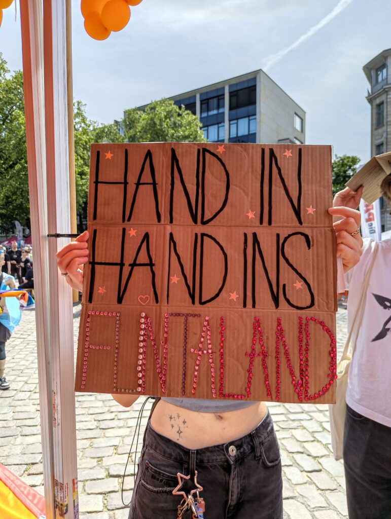 Eine Person hält ein Schild vor ihren Körper. Auf dem Schild in Großbuchstaben der Text »Hand in Hand ins Flintaland«