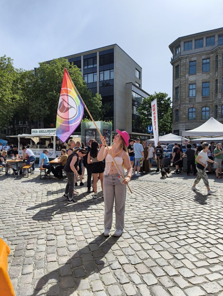 Kyra steht mit einem pinken Hut und einer Regenbogen-Flagge in den Händen auf einem Platz mit Kopfsteinpflaster. Sie schaut in Richtung Flagge. Im Hintergrund Besucher der Stände.