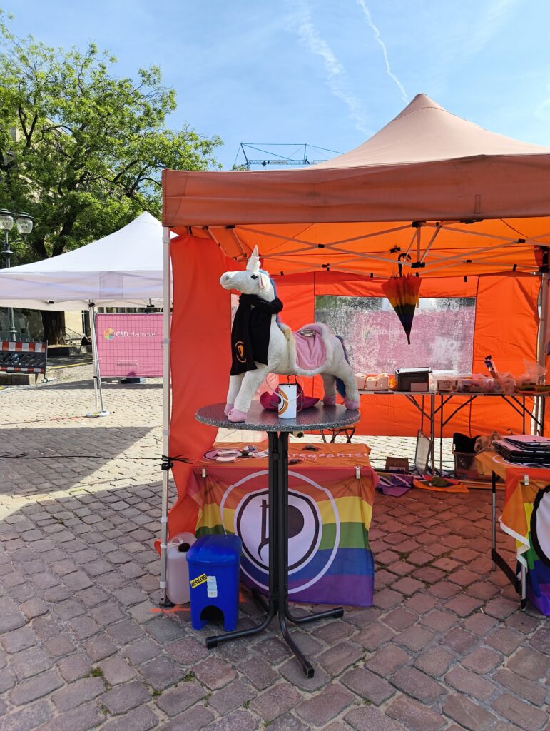 Vor einem orangen Pavillion befindet sich ein runder Stehtisch. Auf dem Stehtisch ein Einhorn mit einem schwarzen Schal der Piratenpartei. An den Tischen des Pavillions Regenbogenflaggen mit Piratenlogo in der Mitte.