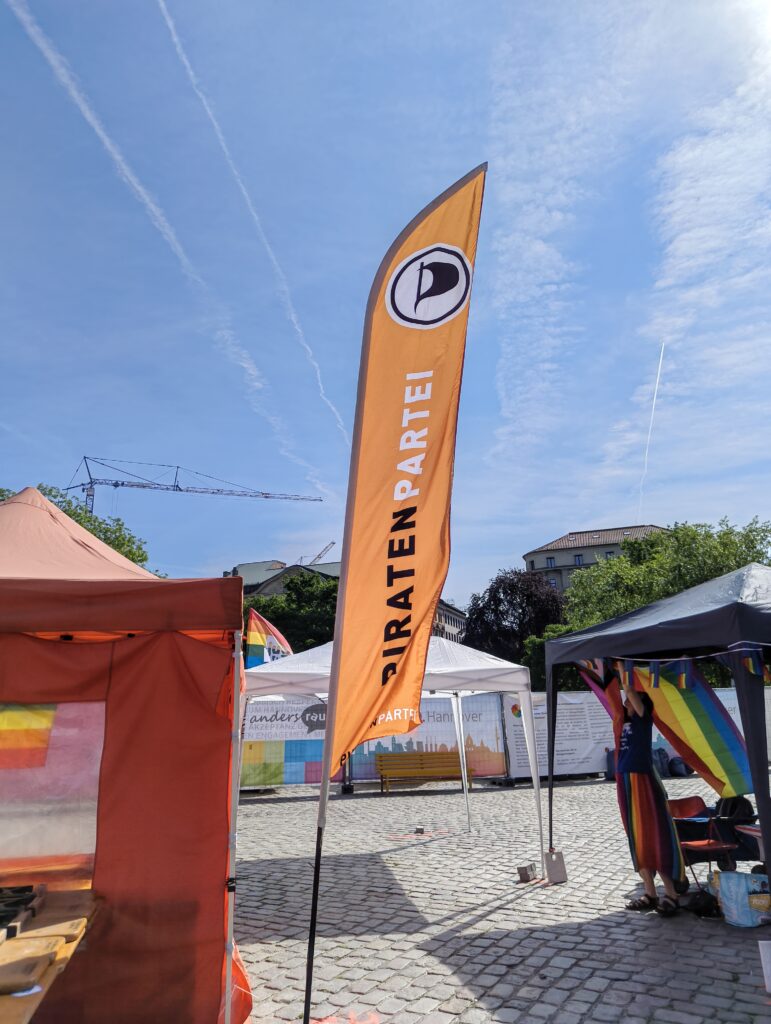 Eine Beach-Flagge mit dem Log der Piratenpartei (Deutschland) steht seitlich neben einem orangen Pavillion.
