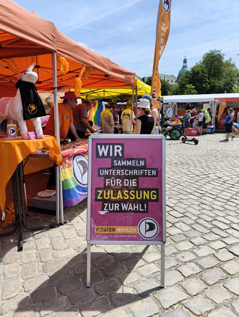 Zwei Personen im Gespräch mit Thomas an unserem Stand. Im Vordergrund unser Aufsteller mit dem Hinweis auf unsere Unterschriftensammlung
