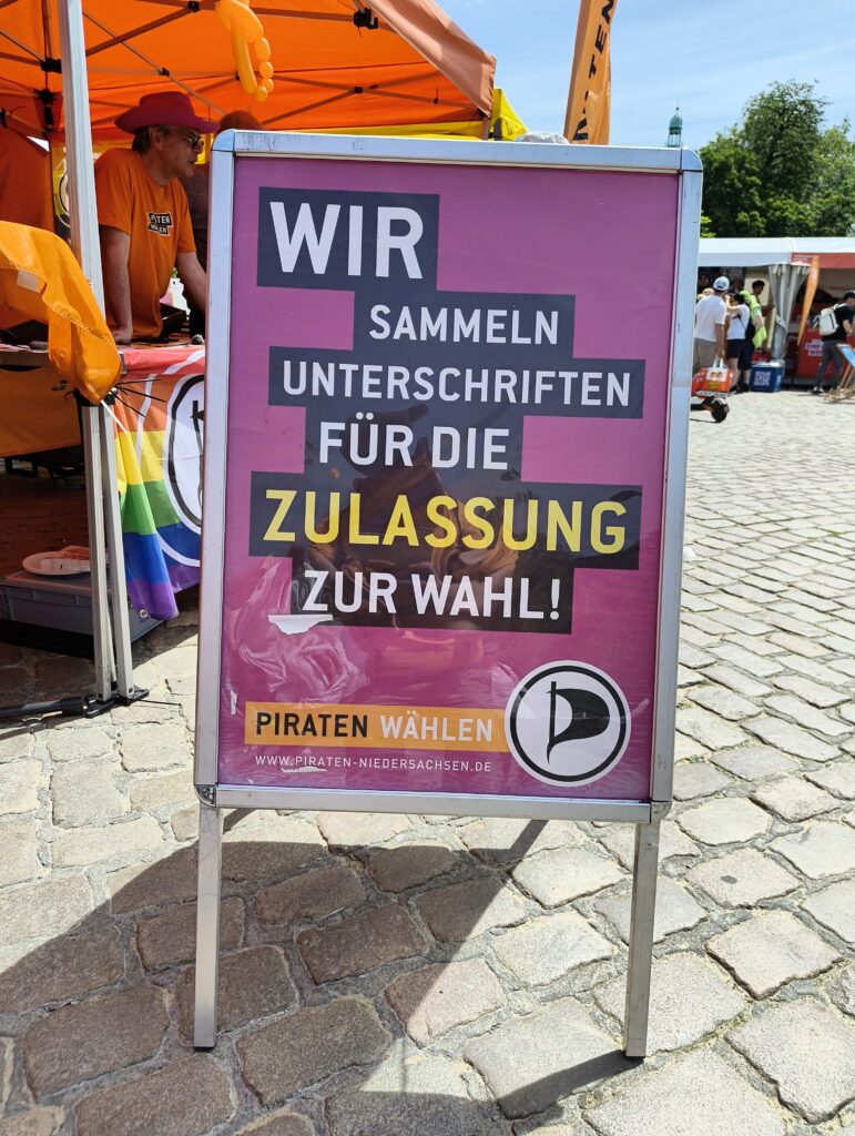 Unser Aufsteller in der Bildmitte: »Wir sammeln Unterschriften für die Zulassung zur Wahl« Darunter der Aufruf »Piraten wählen« und das Piraten-Logo. Im Hintergrund unser Stand.