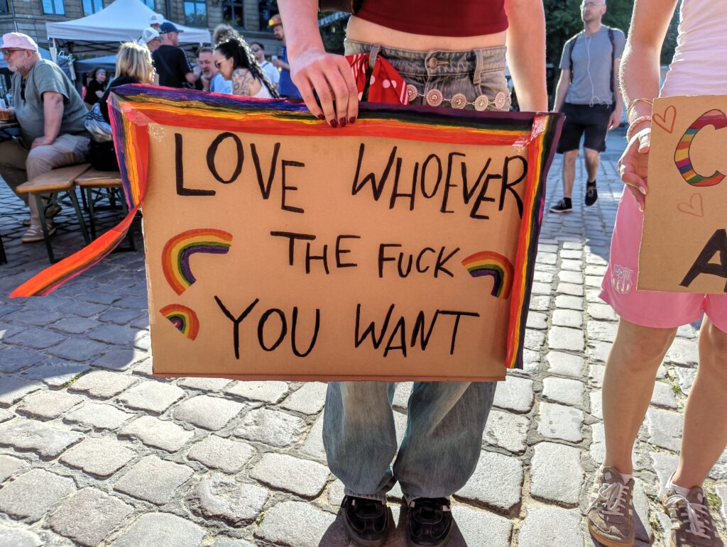 Eine Person hält ein selbst gemachtes Schild vor ihre Beine. Der Text darauf lautet: »Love whoever the fuck you want« Neben dem Text mehrere Regenbögen.