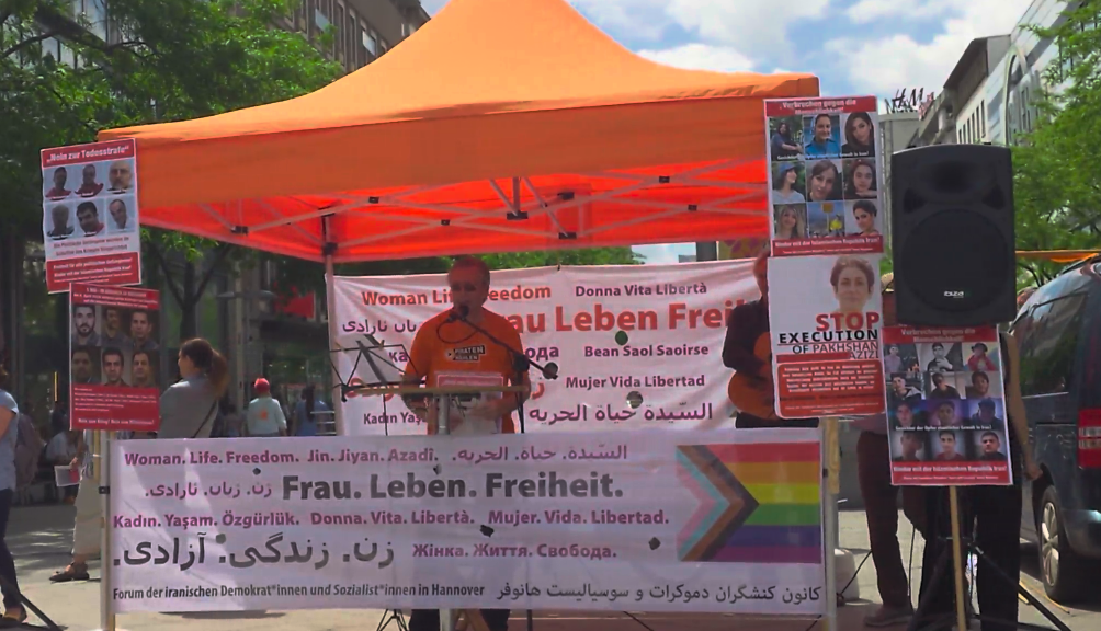 Thomas Ganskow am Mikrofon unter einem orangen Baldachin. Vor und hinter ihm Banner mit den Worten "Frau. Leben. Freiheit." in verschiedenen Sprachen. Rechts und links Lautsprecher auf Ständern.