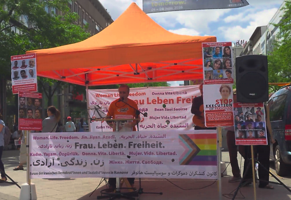 Thomas Ganskow am Mikrofon unter einem orangen Baldachin. Vor und hinter ihm Banner mit den Worten "Frau. Leben. Freiheit." in verschiedenen Sprachen. Rechts und links Lautsprecher auf Ständern.