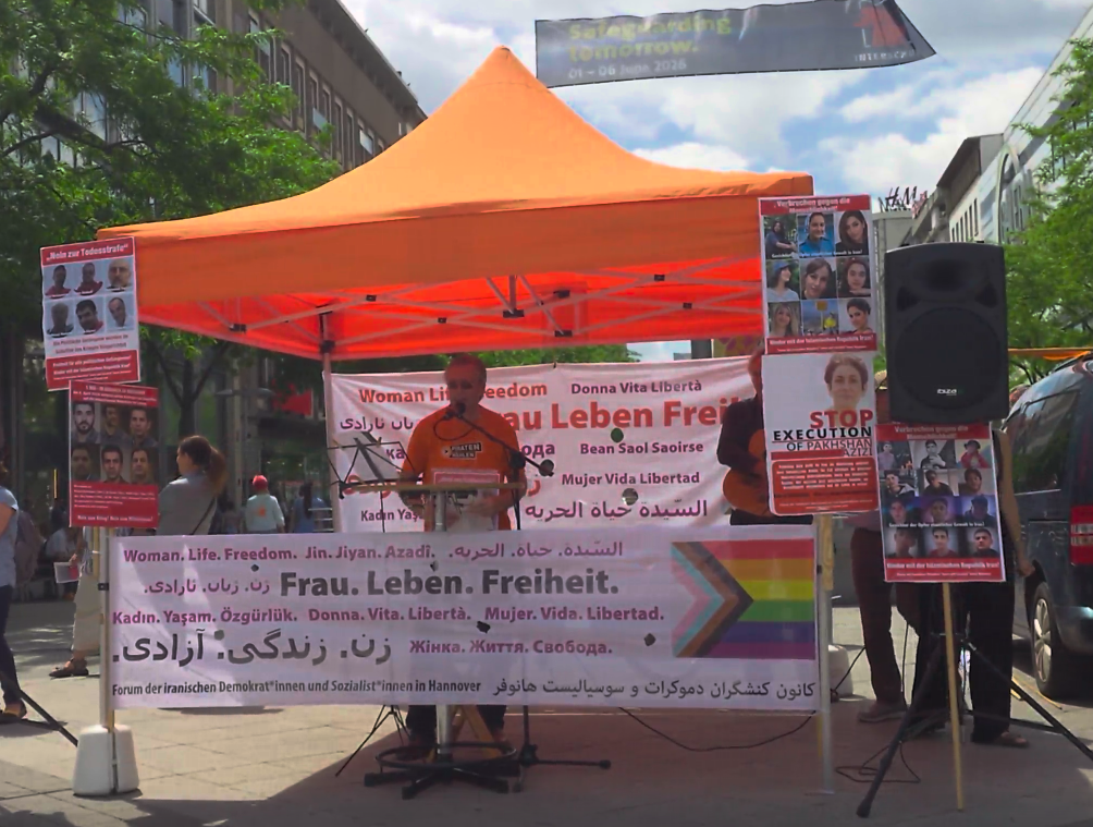Thomas Ganskow am Mikrofon unter einem orangen Baldachin. Vor und hinter ihm Banner mit den Worten "Frau. Leben. Freiheit." in verschiedenen Sprachen. Rechts und links Lautsprecher auf Ständern.