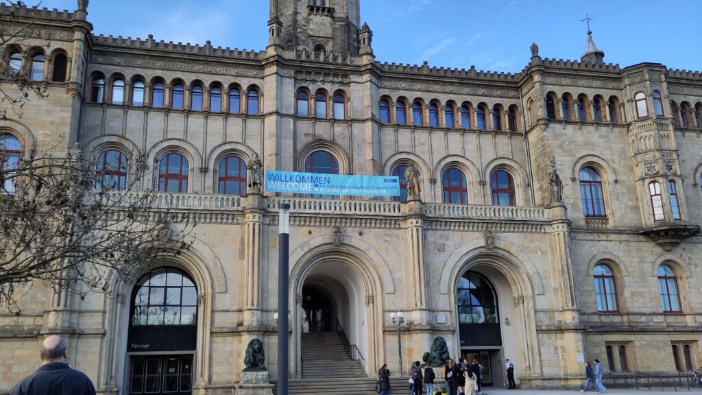 Blick auf das Hauptportal der Universität Hannover. Eine Gruppe hat sich am Fuss der Treppe eingefunden. Ein Transparent ist auf der Brüstung aufgespannt; der Text darauf lautet: „Willkommen an der Leibniz Universität Hannover“