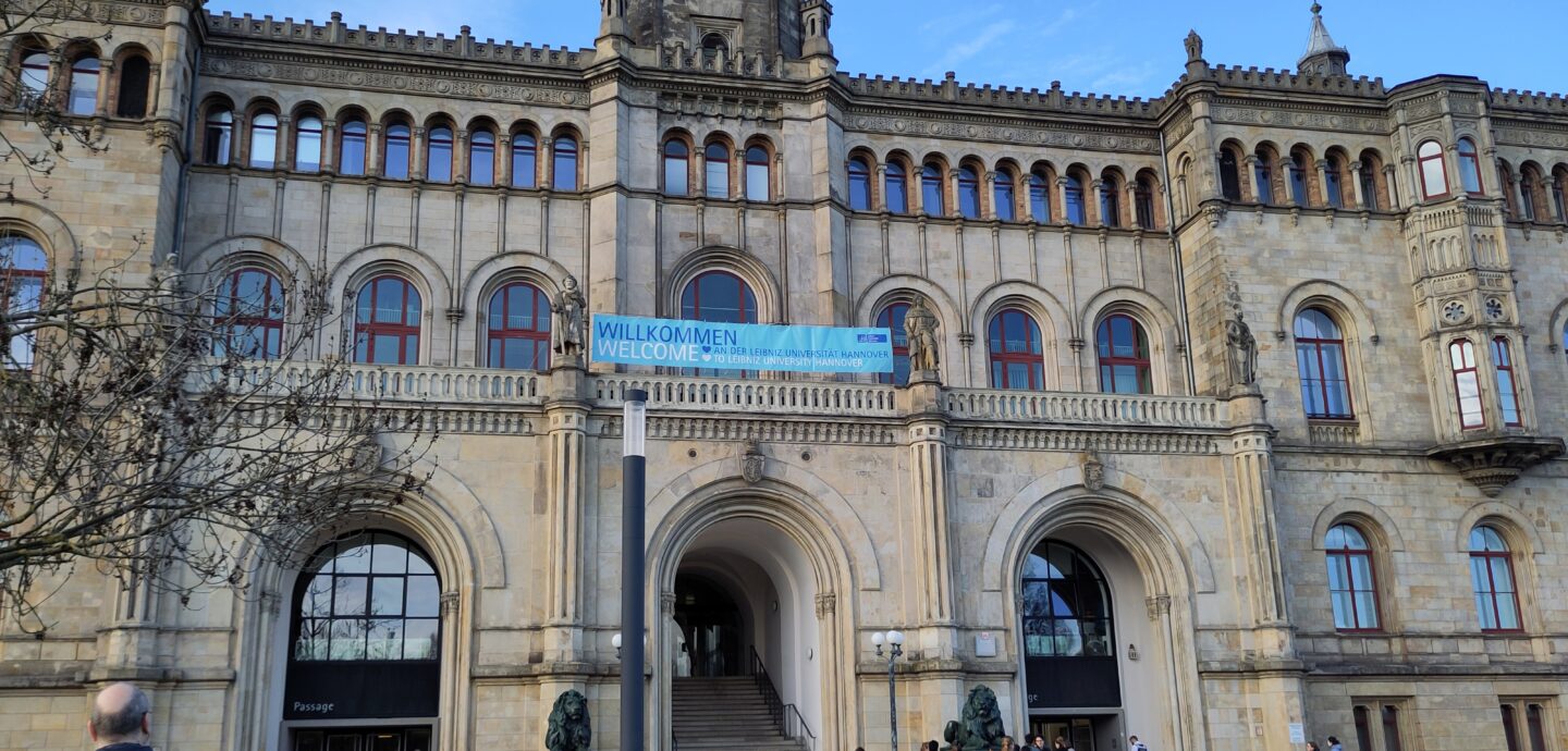 Blick auf das Hauptportal der Universität Hannover. Eine Gruppe hat sich am Fuss der Treppe eingefunden. Ein Transparent ist auf der Brüstung aufgespannt; der Text darauf lautet: „Willkommen an der Leibniz Universität Hannover“