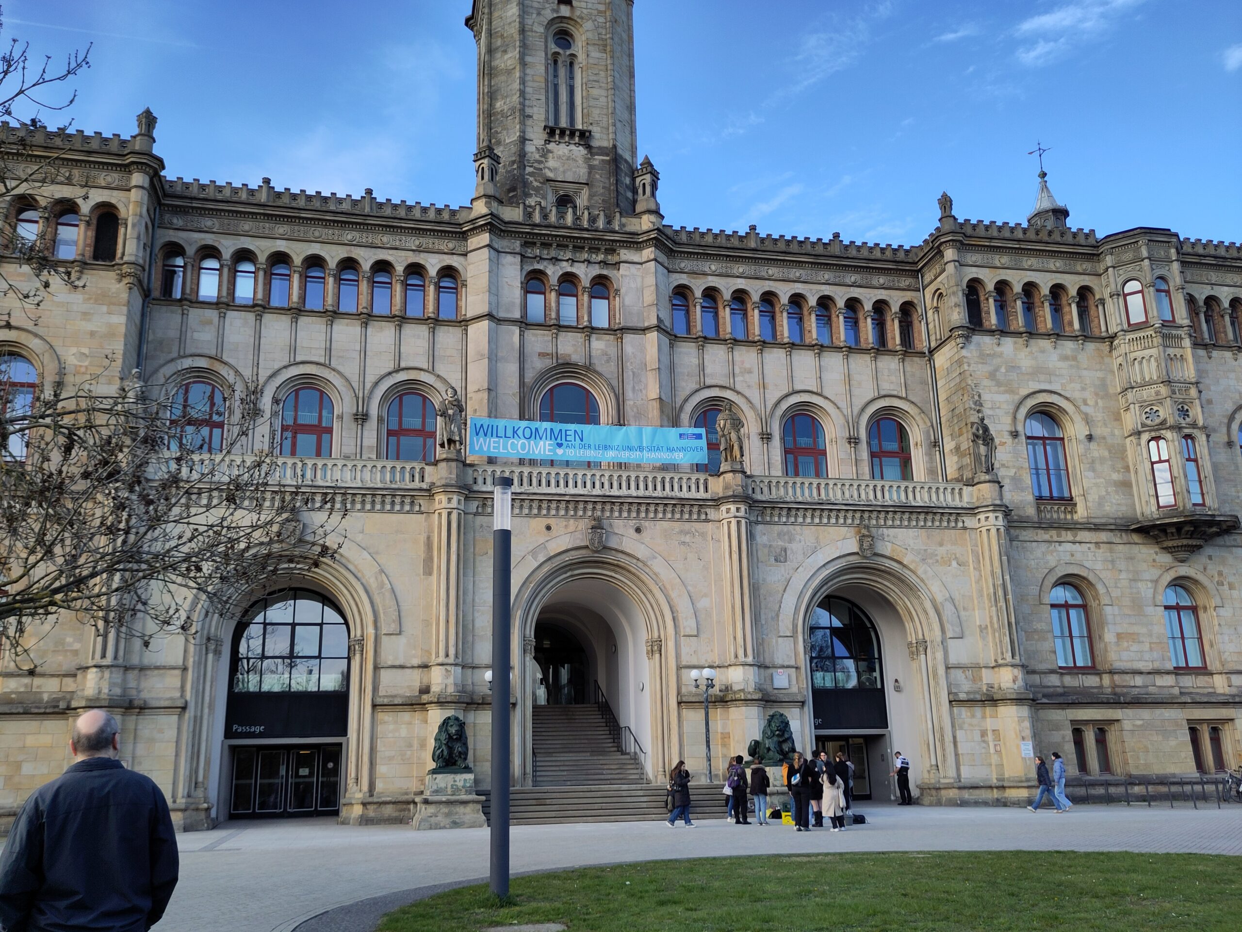 Blick auf das Hauptportal der Universität Hannover. Eine Gruppe hat sich am Fuss der Treppe eingefunden. Ein Transparent ist auf der Brüstung aufgespannt; der Text darauf lautet: „Willkommen an der Leibniz Universität Hannover“